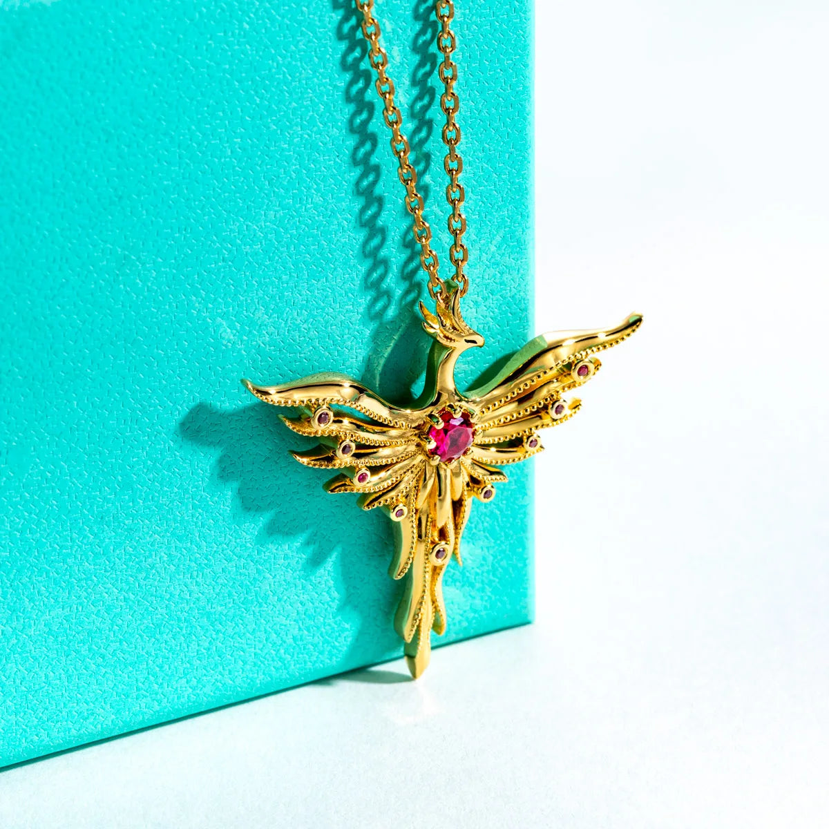 Gold bird-shaped necklace with a red gemstone on a turquoise background
